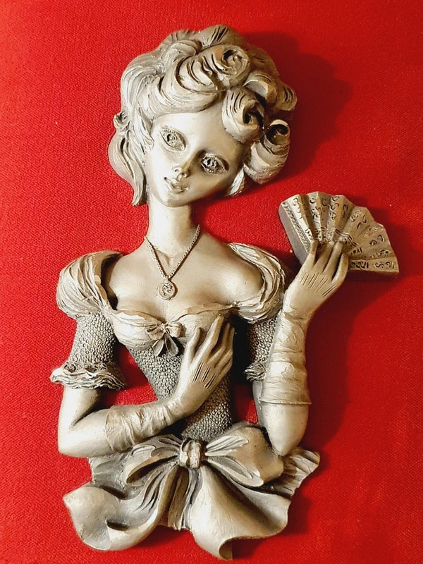 Une mise en scène de style rétro-romantique montrant un coin lecture chaleureux. Au centre, un petit tableau en bas-relief représentant une femme élégante à l'éventail est accroché sur un support en velours rouge, fixé à un mur en briques de bois.
