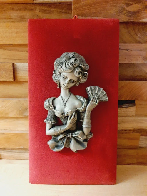 Une mise en scène de style rétro-romantique montrant un coin lecture chaleureux. Au centre, un petit tableau en bas-relief représentant une femme élégante à l'éventail est accroché sur un support en velours rouge, fixé à un mur en briques de bois.