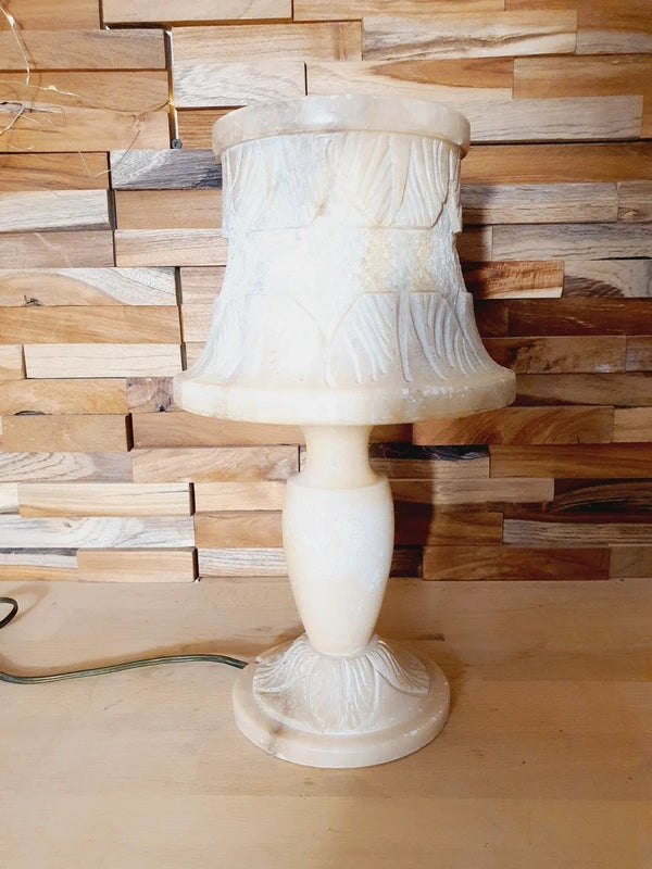 Lampe à poser italienne en albâtre de Volterra éteinte, posée sur un meuble en bois devant un mur de parement en bois. pierre blanche et sculptée créant une atmosphère cosy chic et chaleureuse.