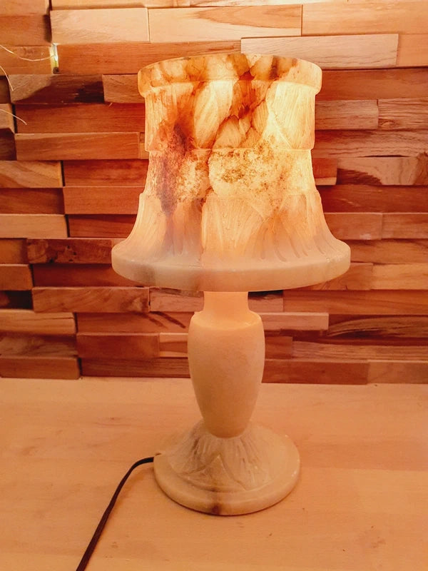 Lampe à poser italienne en albâtre de Volterra allumée, posée sur un meuble en bois devant un mur de parement en bois. La lumière révèle les veines naturelles et organiques de la pierre, créant une atmosphère cosy chic et chaleureuse.