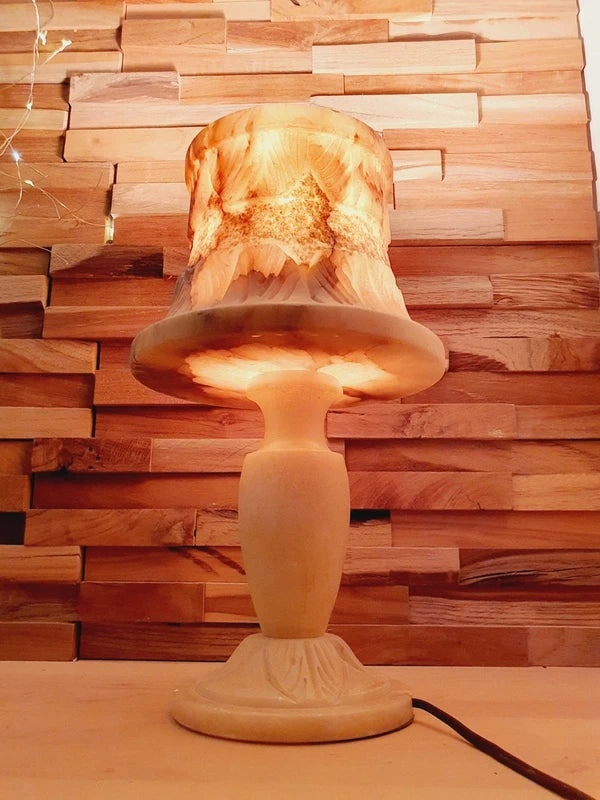 Lampe à poser italienne en albâtre de Volterra allumée, posée sur un meuble en bois devant un mur de parement en bois. La lumière révèle les veines naturelles et organiques de la pierre, créant une atmosphère cosy chic et chaleureuse.