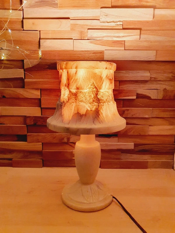 Lampe à poser italienne en albâtre de Volterra allumée, posée sur un meuble en bois devant un mur de parement en bois. La lumière révèle les veines naturelles et organiques de la pierre, créant une atmosphère cosy chic et chaleureuse.