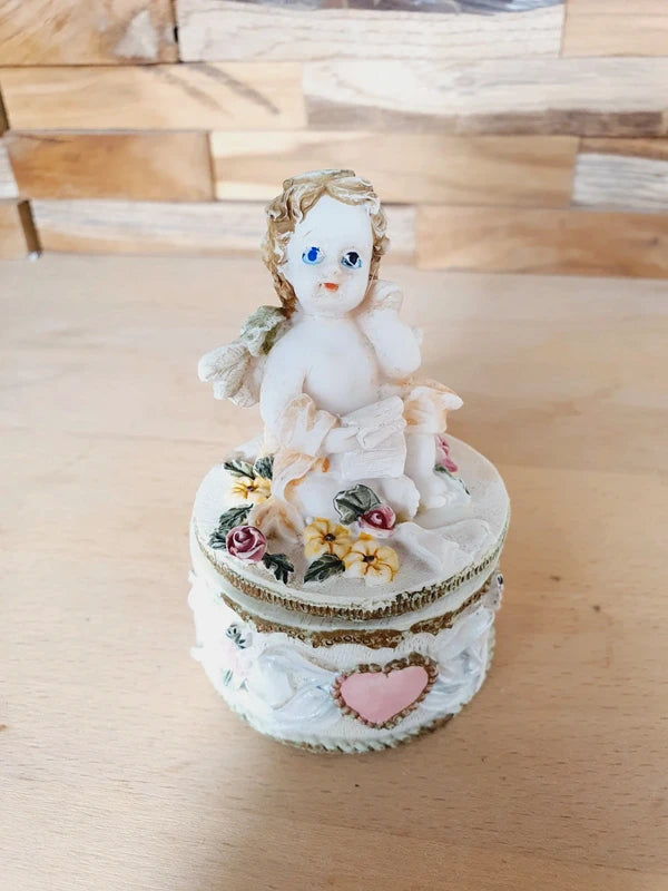Boîte ronde en céramique blanche avec couvercle surmonté d'une figurine d'ange tenant une lettre, ornée de fleurs rouges et jaunes et d'un cœur rose en relief.