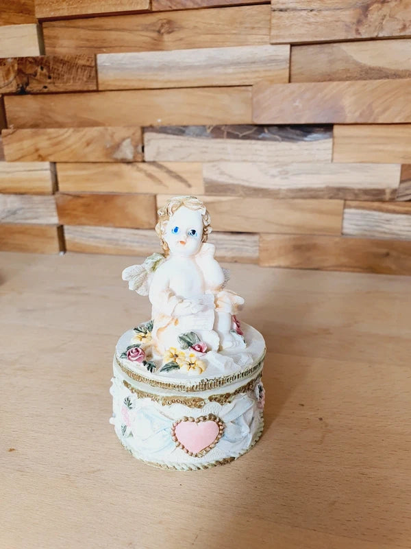 Boîte ronde en céramique blanche avec couvercle surmonté d'une figurine d'ange tenant une lettre, ornée de fleurs rouges et jaunes et d'un cœur rose en relief.