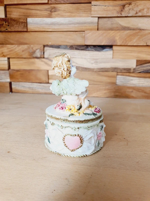 Boîte ronde en céramique blanche avec couvercle surmonté d'une figurine d'ange tenant une lettre, ornée de fleurs rouges et jaunes et d'un cœur rose en relief.