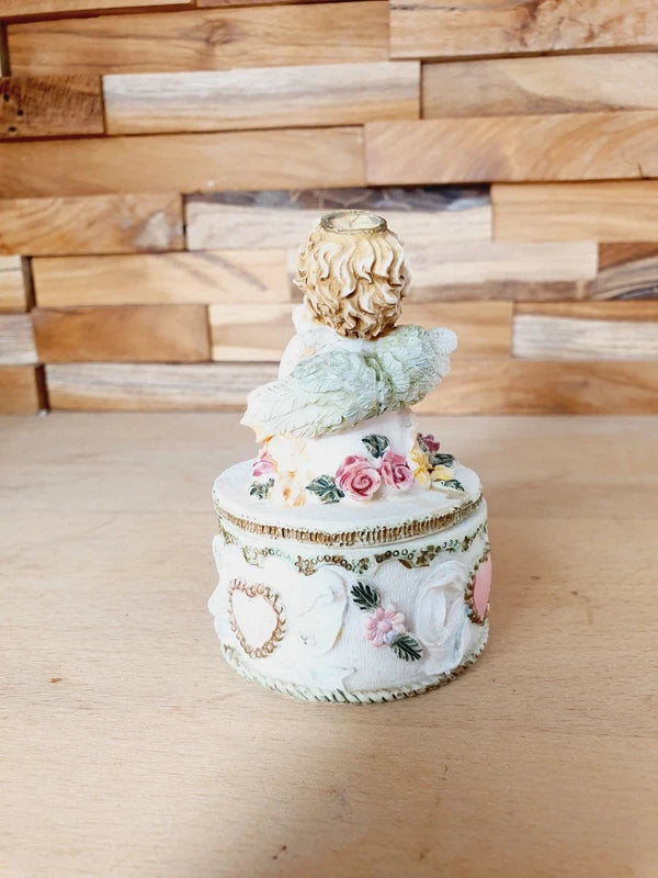 Boîte ronde en céramique blanche avec couvercle surmonté d'une figurine d'ange tenant une lettre, ornée de fleurs rouges et jaunes et d'un cœur rose en relief.