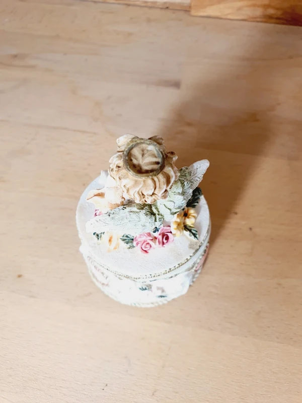 Boîte ronde en céramique blanche avec couvercle surmonté d'une figurine d'ange tenant une lettre, ornée de fleurs rouges et jaunes et d'un cœur rose en relief.
