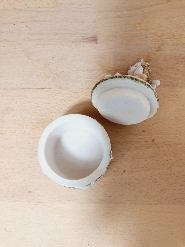 Boîte ronde en céramique blanche avec couvercle surmonté d'une figurine d'ange tenant une lettre, ornée de fleurs rouges et jaunes et d'un cœur rose en relief.