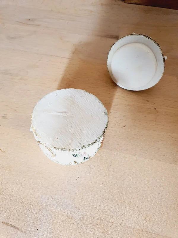 Boîte ronde en céramique blanche avec couvercle surmonté d'une figurine d'ange tenant une lettre, ornée de fleurs rouges et jaunes et d'un cœur rose en relief.