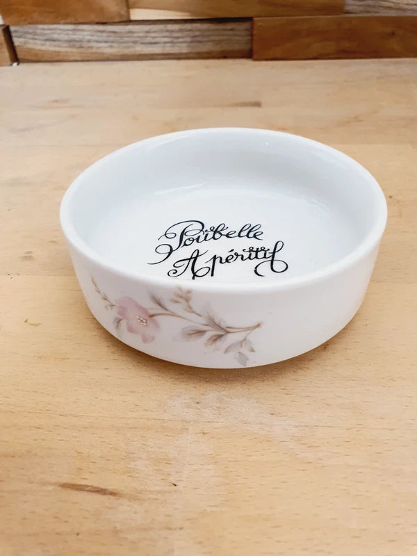 Petite coupelle ronde en porcelaine blanche brillante avec l'inscription noire "Poubelle Apéritif" à l'intérieur et de fines fleurs colorées sur le bord extérieur.