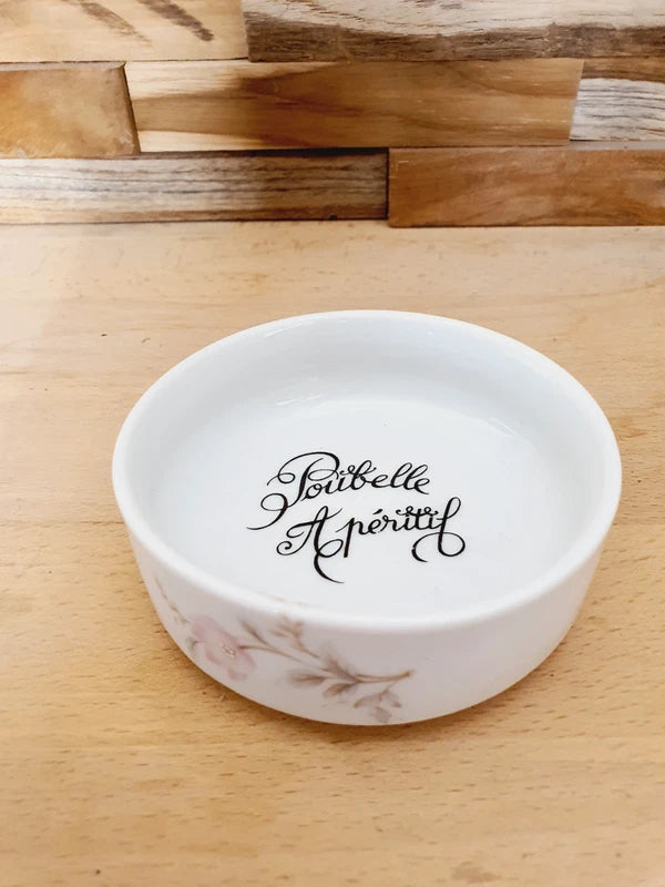 Petite coupelle ronde en porcelaine blanche brillante avec l'inscription noire "Poubelle Apéritif" à l'intérieur et de fines fleurs colorées sur le bord extérieur.