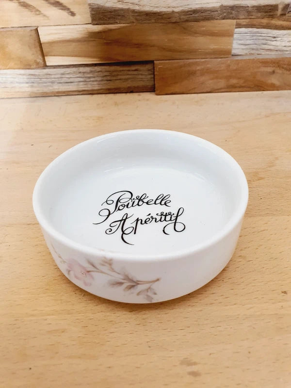 Petite coupelle ronde en porcelaine blanche brillante avec l'inscription noire "Poubelle Apéritif" à l'intérieur et de fines fleurs colorées sur le bord extérieur.
