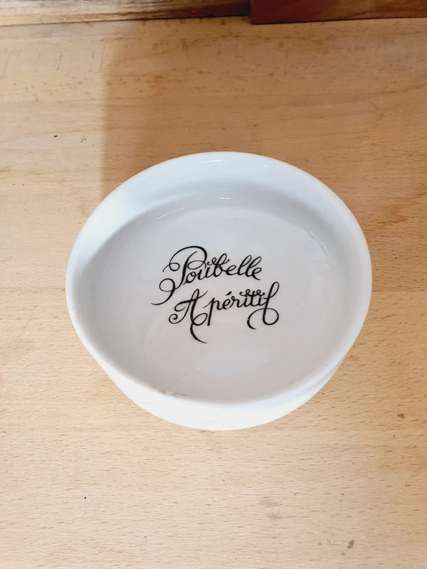 Petite coupelle ronde en porcelaine blanche brillante avec l'inscription noire "Poubelle Apéritif" à l'intérieur et de fines fleurs colorées sur le bord extérieur.