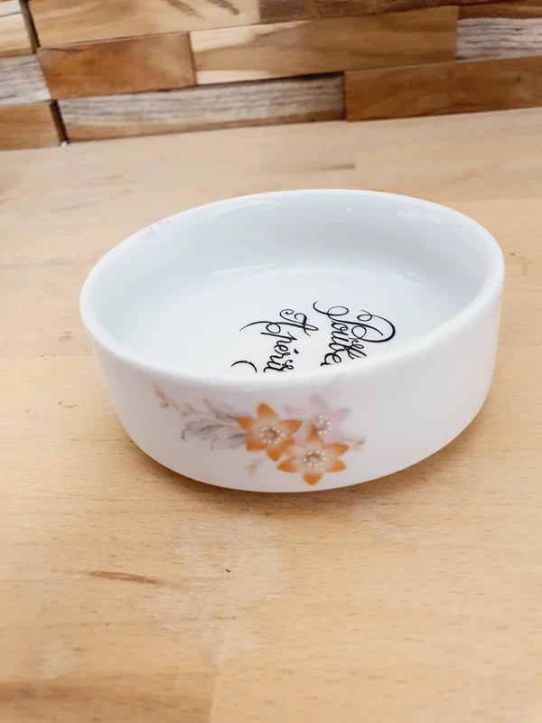 Petite coupelle ronde en porcelaine blanche brillante avec l'inscription noire "Poubelle Apéritif" à l'intérieur et de fines fleurs colorées sur le bord extérieur.