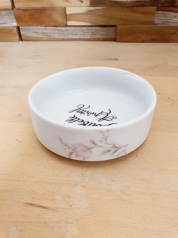 Petite coupelle ronde en porcelaine blanche brillante avec l'inscription noire "Poubelle Apéritif" à l'intérieur et de fines fleurs colorées sur le bord extérieur.