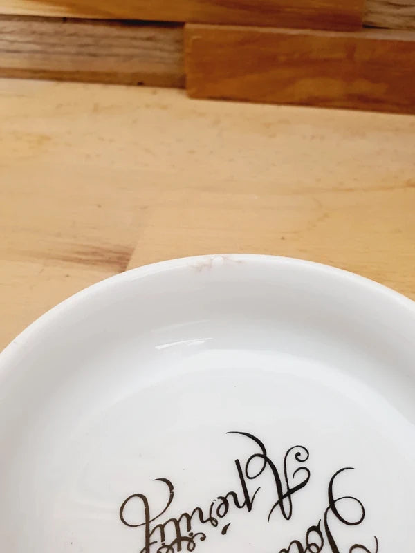 Petite coupelle ronde en porcelaine blanche brillante avec l'inscription noire "Poubelle Apéritif" à l'intérieur et de fines fleurs colorées sur le bord extérieur.