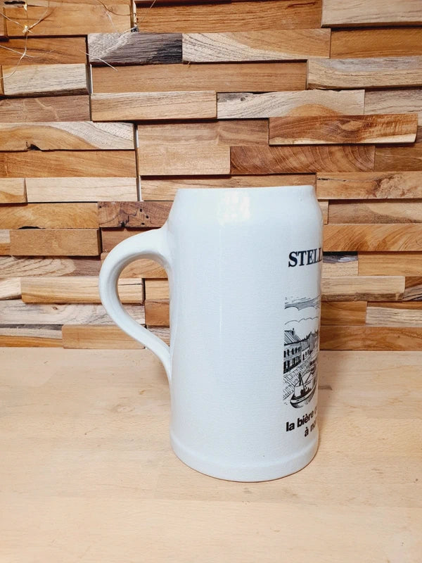 Grande choppe à bière en grès beige portant le logo Stella Artois et l'inscription noire "la bière qui donne du relief à notre plat pays" avec une illustration de péniche.