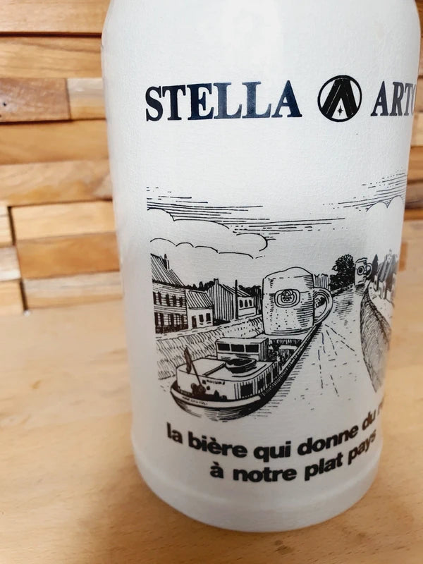 Grande choppe à bière en grès beige portant le logo Stella Artois et l'inscription noire "la bière qui donne du relief à notre plat pays" avec une illustration de péniche.