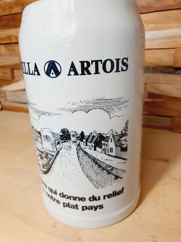 Grande choppe à bière en grès beige portant le logo Stella Artois et l'inscription noire "la bière qui donne du relief à notre plat pays" avec une illustration de péniche.