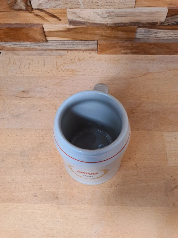 Petite choppe à bière vintage Stella Artois en grès gris avec logo rouge et or, format 25 cl, posée sur un fond en bois.