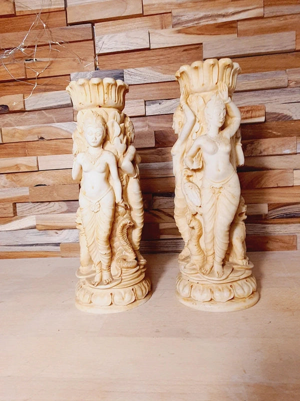 Paire bougeoirs sculptés résine ivoire au décor riche de déesses et d'animaux, sur un fond de bois clair.