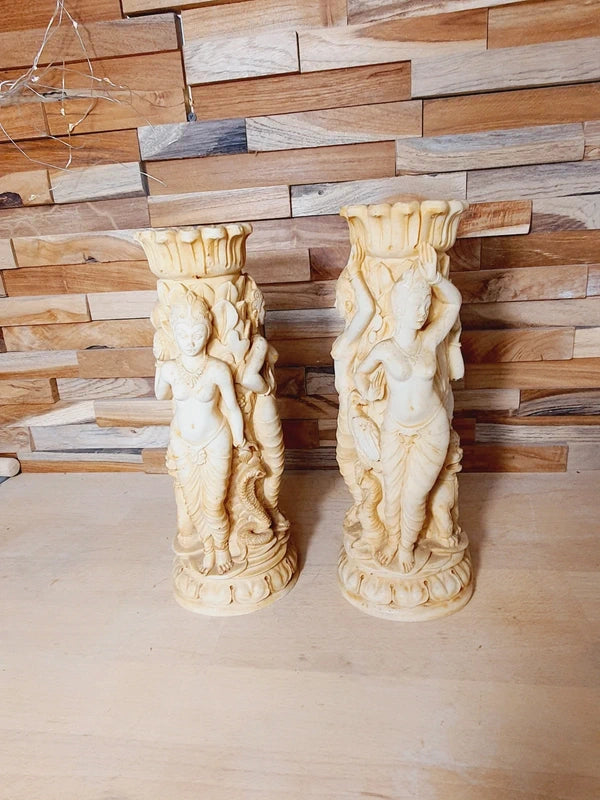 Paire bougeoirs sculptés résine ivoire au décor riche de déesses et d'animaux, sur un fond de bois clair.
