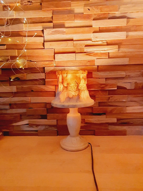 Lampe à poser italienne en albâtre de Volterra allumée, posée sur un meuble en bois devant un mur de parement en bois. La lumière révèle les veines naturelles et organiques de la pierre, créant une atmosphère cosy chic et chaleureuse.