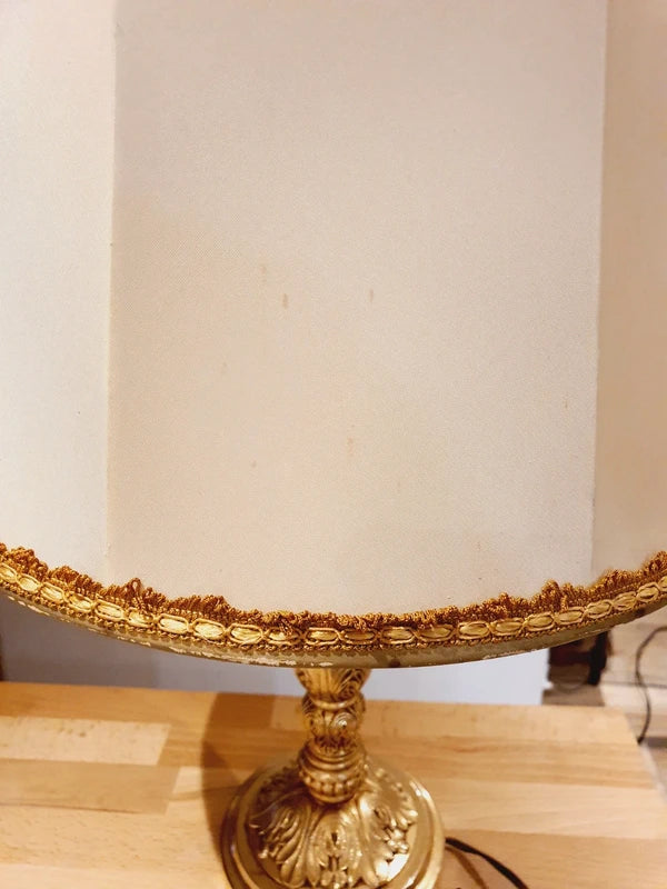 Lampe de table de style Regency & Art Français années 50-60. Hauteur 55,5 cm. Elle possède un pied en laiton sculpté combinant une base ornée. Son grand abat-jour en tissu de couleur crème est bordé de galons décoratifs. 