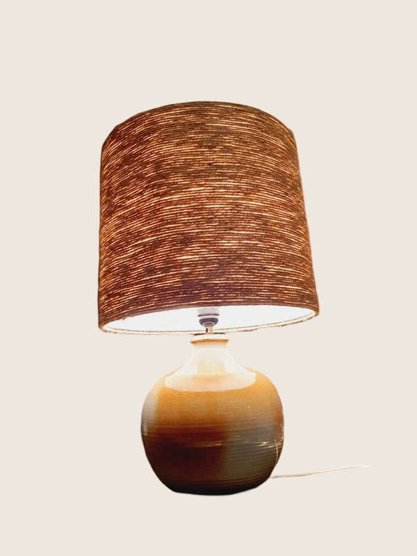 Une lampe vintage imposante composée d'un pied en grès français beige moucheté et d'un grand abat-jour en toile de jute tressée, diffusant une lumière chaude dans un intérieur à la décoration naturelle.
