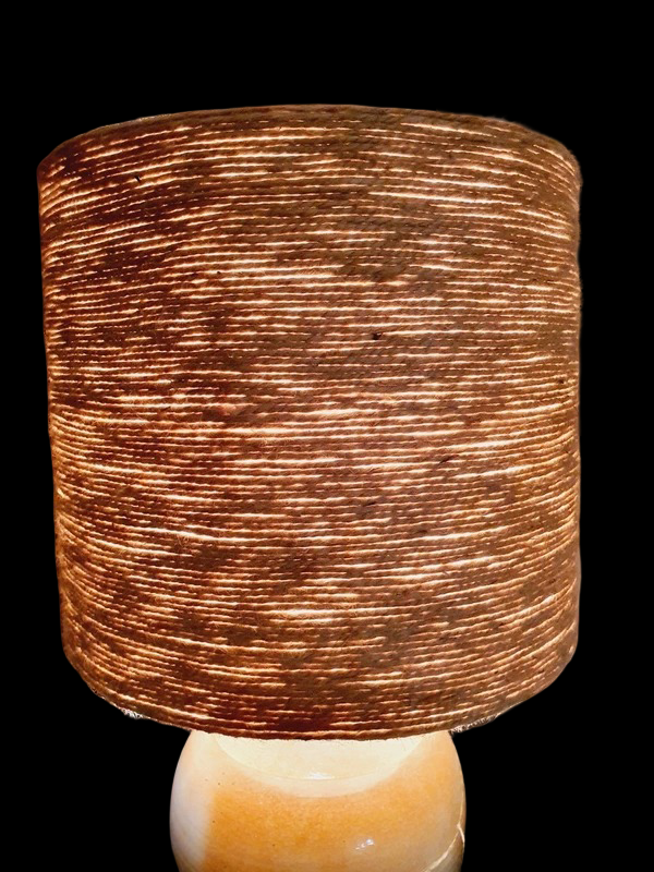Une lampe vintage imposante composée d'un pied en grès français beige moucheté et d'un grand abat-jour en toile de jute tressée, diffusant une lumière chaude dans un intérieur à la décoration naturelle.