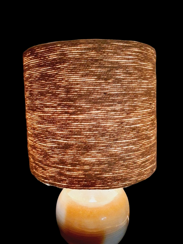 Une lampe vintage imposante composée d'un pied en grès français beige moucheté et d'un grand abat-jour en toile de jute tressée, diffusant une lumière chaude dans un intérieur à la décoration naturelle.