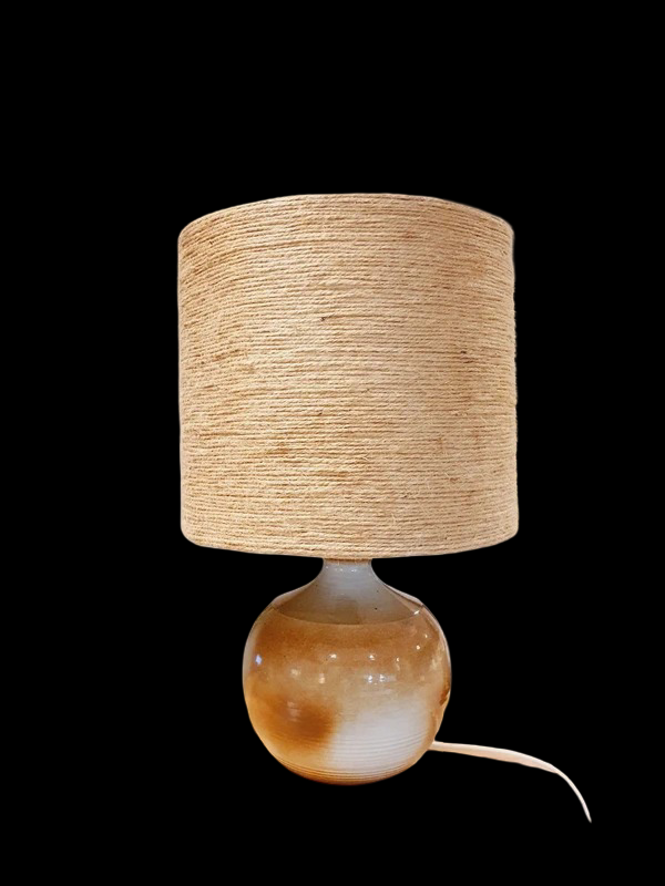 Une lampe vintage imposante composée d'un pied en grès français beige moucheté et d'un grand abat-jour en toile de jute tressée, diffusant une lumière chaude dans un intérieur à la décoration naturelle.