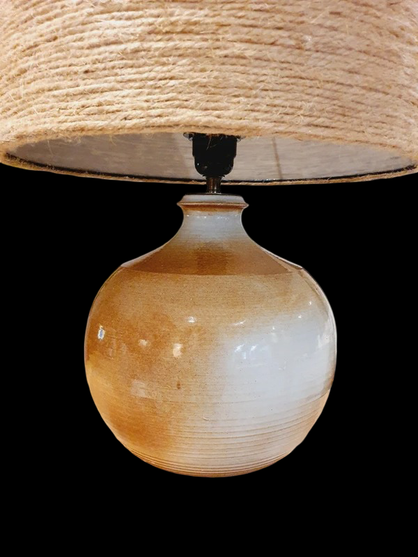 Une lampe vintage imposante composée d'un pied en grès français beige moucheté et d'un grand abat-jour en toile de jute tressée, diffusant une lumière chaude dans un intérieur à la décoration naturelle.