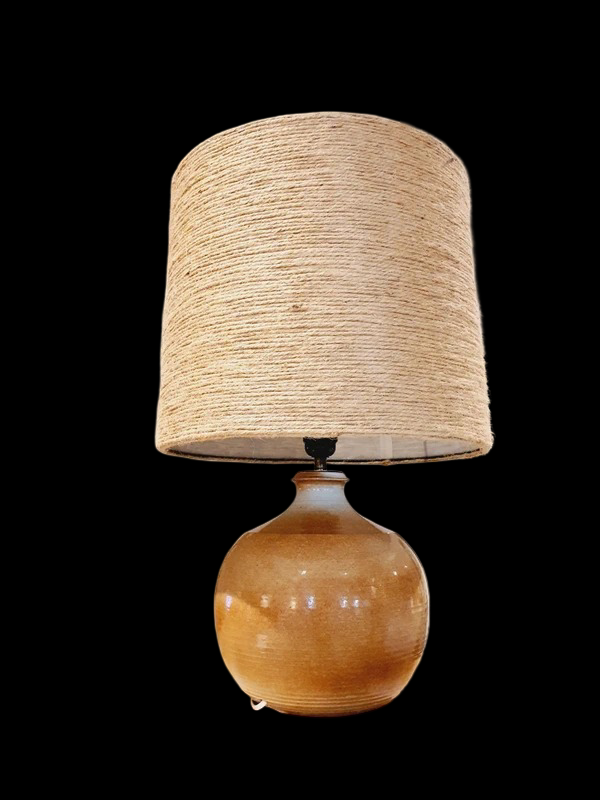 Une lampe vintage imposante composée d'un pied en grès français beige moucheté et d'un grand abat-jour en toile de jute tressée, diffusant une lumière chaude dans un intérieur à la décoration naturelle.