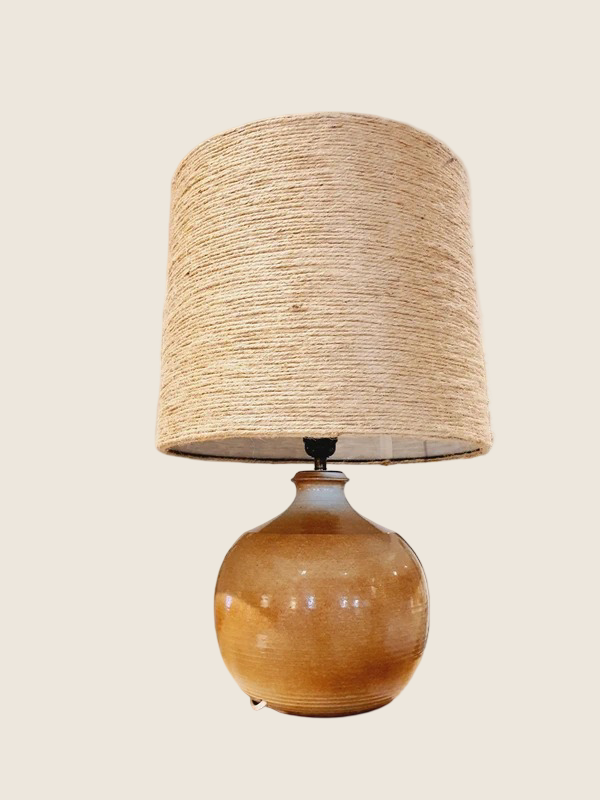 Une lampe vintage imposante composée d'un pied en grès français beige moucheté et d'un grand abat-jour en toile de jute tressée, diffusant une lumière chaude dans un intérieur à la décoration naturelle.
