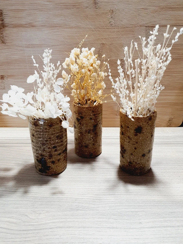 Lot de 3 petits gobelets cylindriques en grès pyrité vintage fait main des années 1970 aux nuances ocres et brunes mouchetées avec une finition vernissée brillante, mis en scène séparément et debout garnis de petits bouquets de fleurs séchées blanches et crèmes.