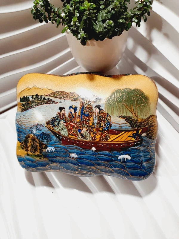 Boîte à bijoux rectangulaire festonnée en porcelaine vintage style Satsuma des années 1970 avec un couvercle illustré d'une scène traditionnelle de femmes et fillette sur une pirogue, richement décorée de fleurs colorées et de détails dorés à l'or fin, montrant le dessous estampillé d'une étiquette rouge Made in China Satsuma.