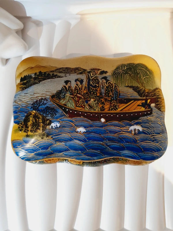 Boîte à bijoux rectangulaire festonnée en porcelaine vintage style Satsuma des années 1970 avec un couvercle illustré d'une scène traditionnelle de femmes et fillette sur une pirogue, richement décorée de fleurs colorées et de détails dorés à l'or fin, montrant le dessous estampillé d'une étiquette rouge Made in China Satsuma.