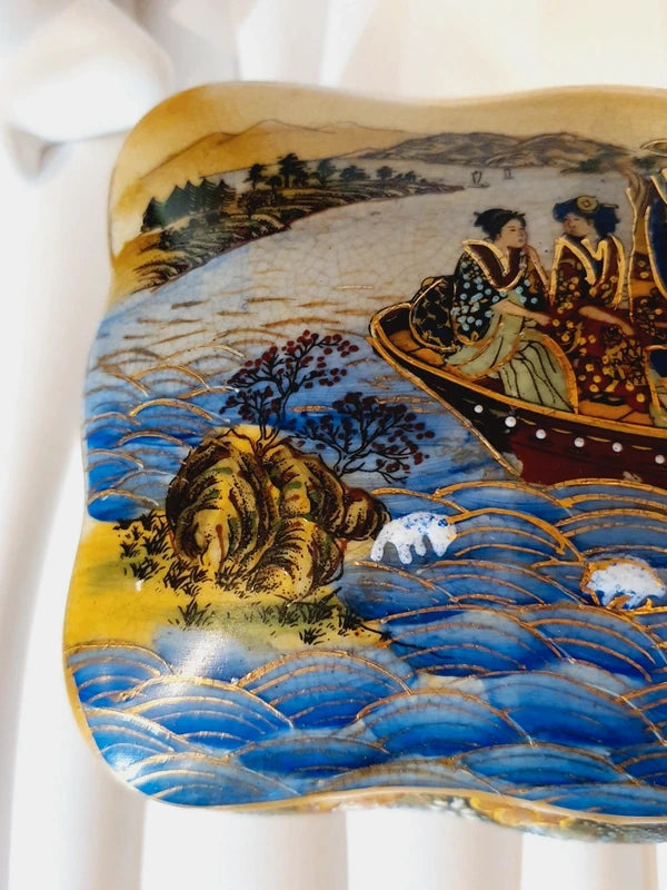 Boîte à bijoux rectangulaire festonnée en porcelaine vintage style Satsuma des années 1970 avec un couvercle illustré d'une scène traditionnelle de femmes et fillette sur une pirogue, richement décorée de fleurs colorées et de détails dorés à l'or fin, montrant le dessous estampillé d'une étiquette rouge Made in China Satsuma.