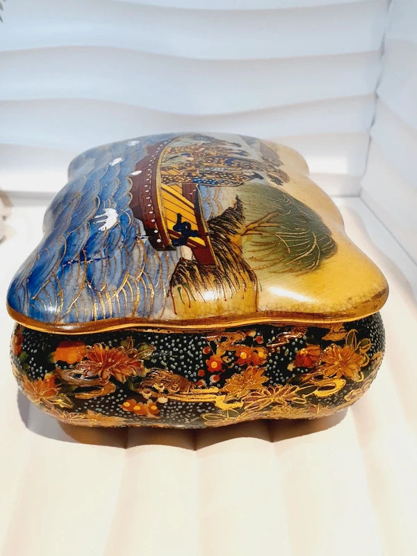 Boîte à bijoux rectangulaire festonnée en porcelaine vintage style Satsuma des années 1970 avec un couvercle illustré d'une scène traditionnelle de femmes et fillette sur une pirogue, richement décorée de fleurs colorées et de détails dorés à l'or fin, montrant le dessous estampillé d'une étiquette rouge Made in China Satsuma.