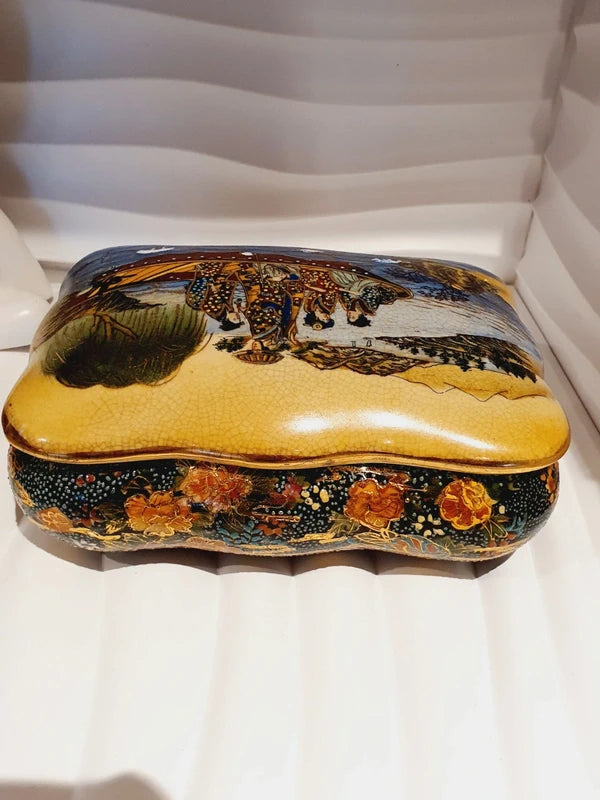 Boîte à bijoux rectangulaire festonnée en porcelaine vintage style Satsuma des années 1970 avec un couvercle illustré d'une scène traditionnelle de femmes et fillette sur une pirogue, richement décorée de fleurs colorées et de détails dorés à l'or fin, montrant le dessous estampillé d'une étiquette rouge Made in China Satsuma.