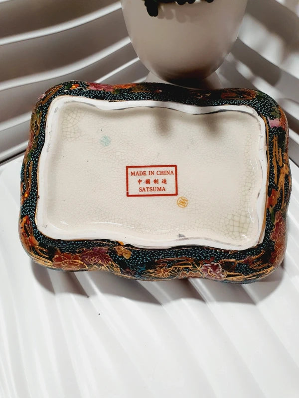 Boîte à bijoux rectangulaire festonnée en porcelaine vintage style Satsuma des années 1970 avec un couvercle illustré d'une scène traditionnelle de femmes et fillette sur une pirogue, richement décorée de fleurs colorées et de détails dorés à l'or fin, montrant le dessous estampillé d'une étiquette rouge Made in China Satsuma.