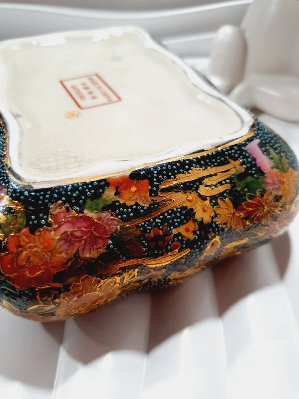 Boîte à bijoux rectangulaire festonnée en porcelaine vintage style Satsuma des années 1970 avec un couvercle illustré d'une scène traditionnelle de femmes et fillette sur une pirogue, richement décorée de fleurs colorées et de détails dorés à l'or fin, montrant le dessous estampillé d'une étiquette rouge Made in China Satsuma.