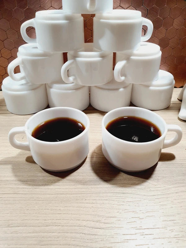 Lot de 12 petites tasses à café empilables en verre opale blanc de lait de la marque Arcopal des années 1970 en parfait état de conservation, présentées d'abord assemblées en pyramide puis au premier plan remplies de café noir fumant.