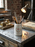 Petite choppe à bière vintage Stella Artois en grès gris avec logo rouge et or, format 25 cl, posée sur un fun bureau style industriel pour ranger stylo, pinceaux, crayons.