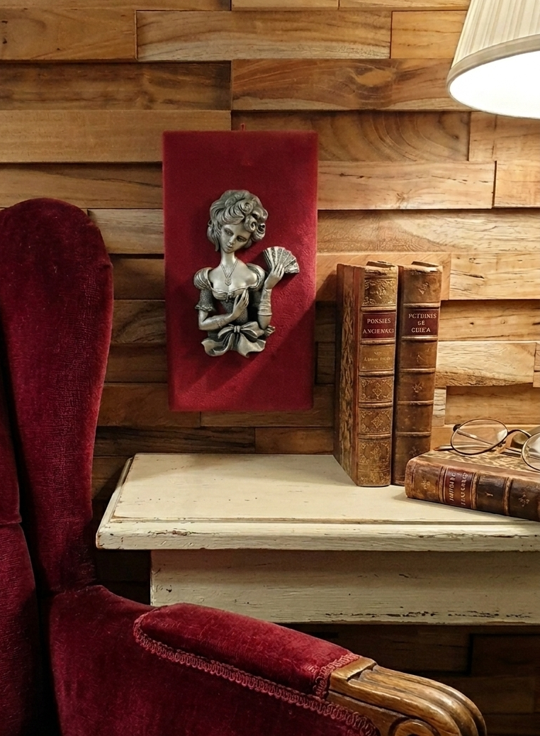 Une mise en scène de style rétro-romantique montrant un coin lecture chaleureux. Au centre, un petit tableau en bas-relief représentant une femme élégante à l'éventail est accroché sur un support en velours rouge, fixé à un mur en briques de bois.