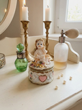 Boîte ronde en céramique blanche avec couvercle surmonté d'une figurine d'ange tenant une lettre, ornée de fleurs rouges et jaunes et d'un cœur rose en relief.